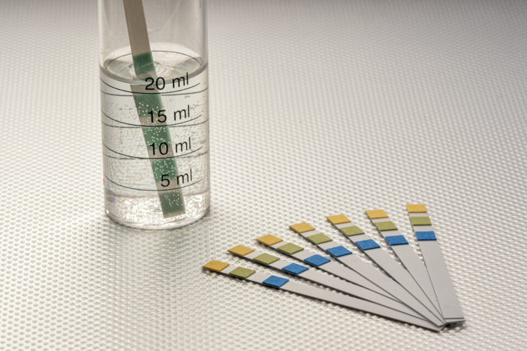 close up of ph analysis strips with a strip inserted in a graduated tube with water, on a rough metal surface