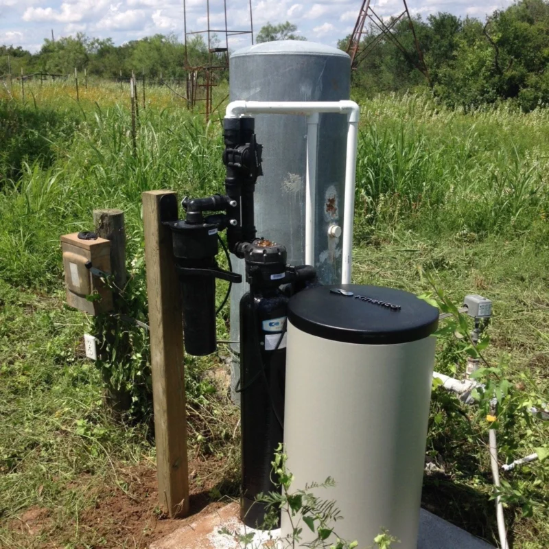 A water well system with a pump, pressure tank, filtration unit, and electrical box sits on a concrete slab in a grassy outdoor area with vegetation and trees in the background.