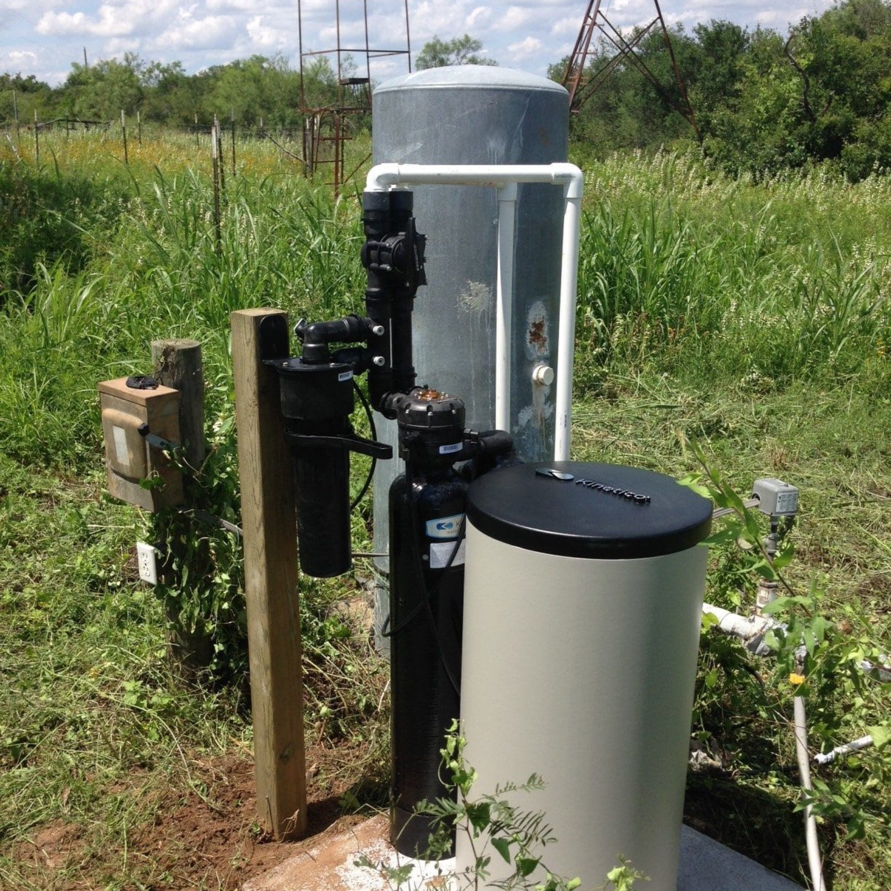 A water well system with a pump, pressure tank, filtration unit, and electrical box sits on a concrete slab in a grassy outdoor area with vegetation and trees in the background.