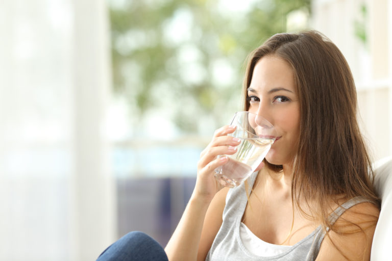 Woman drinking water at home and looking at you sitting on a couch in the living room at home