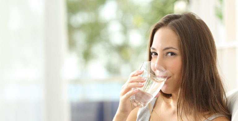 Woman drinking water at home and looking at you sitting on a couch in the living room at home