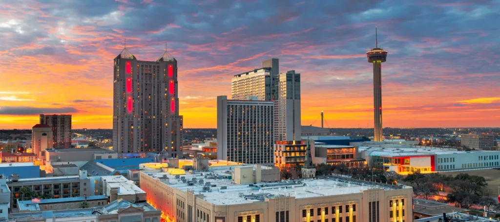 A vibrant city skyline at sunset with tall buildings, a prominent observation tower, and a dramatic sky filled with orange, pink, and blue clouds. Rooftops and streets are lightly illuminated in the foreground.