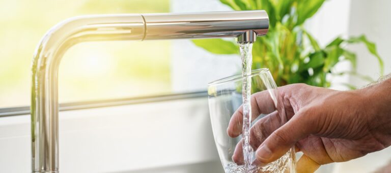 a man filling up a glass of water at his kitchen sink with sunlight shining in through a window in the background