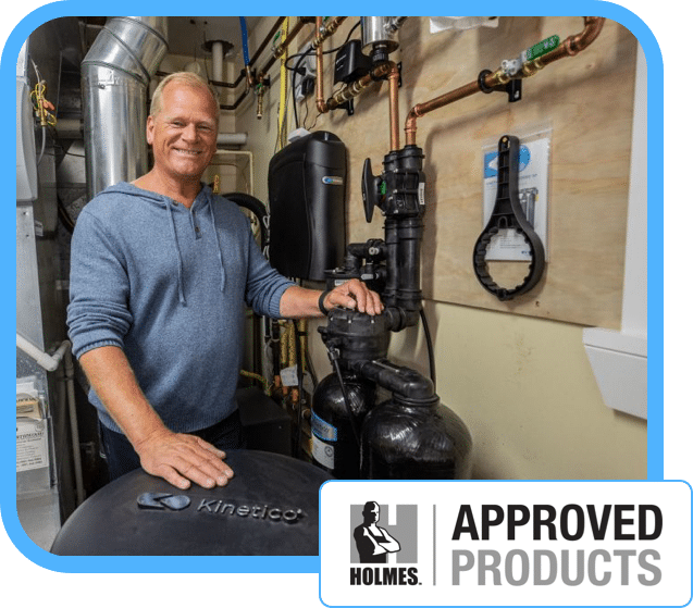 A smiling man in a blue hoodie stands in a utility room beside Kinetico water treatment equipment. The corner features a Holmes Approved Products logo in the bottom right.
