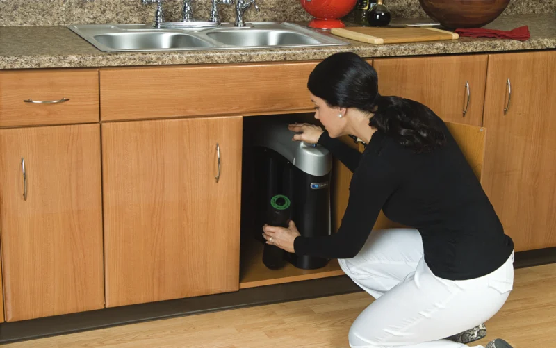 A woman kneels in front of an open kitchen cabinet under a sink, inspecting or adjusting a black and silver appliance installed inside the cabinet.