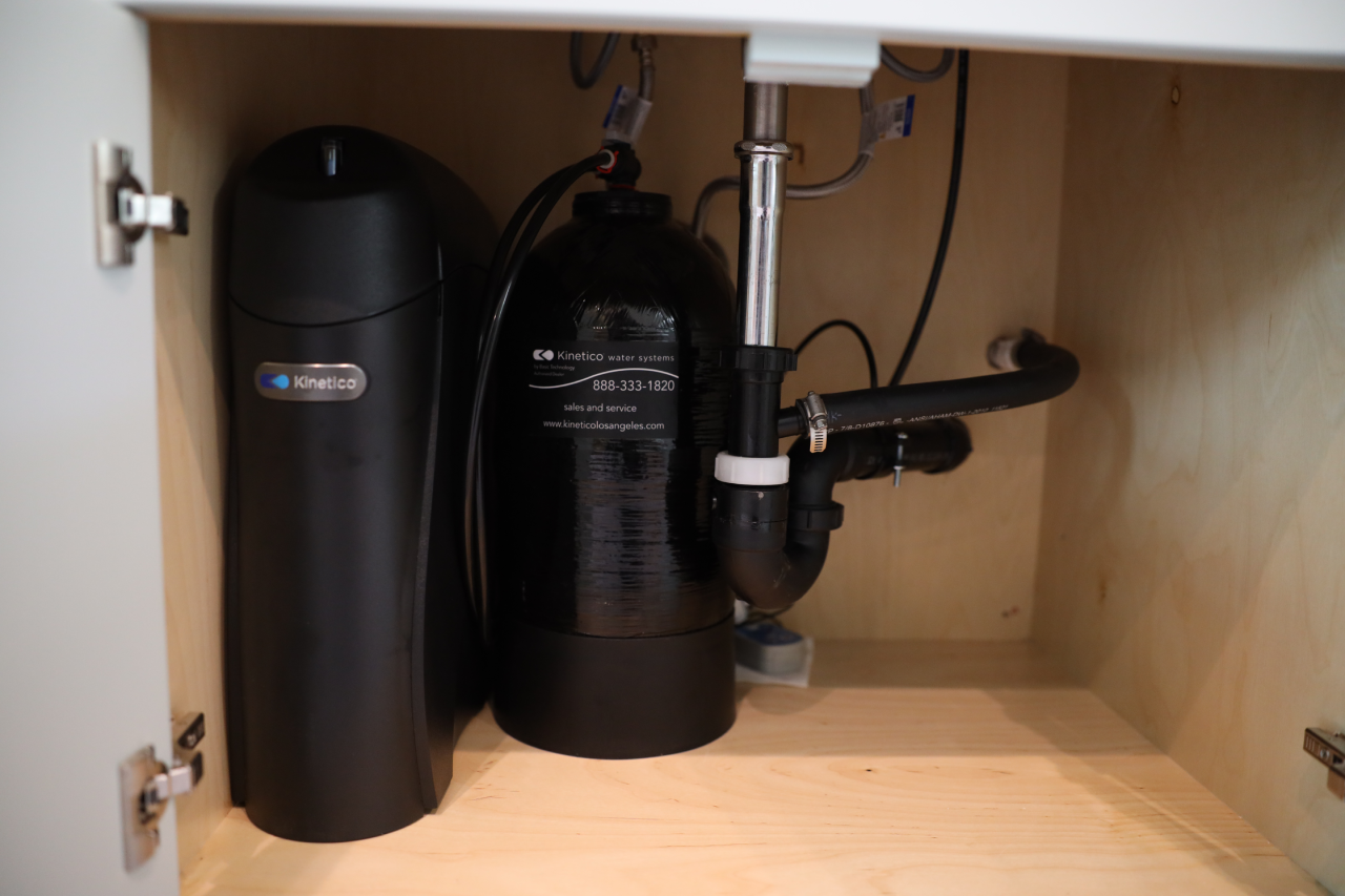 A water filtration system with black cylindrical units and attached pipes is installed under a wooden sink cabinet. The components are labeled “Kinetico,” and plumbing fixtures are visible nearby.