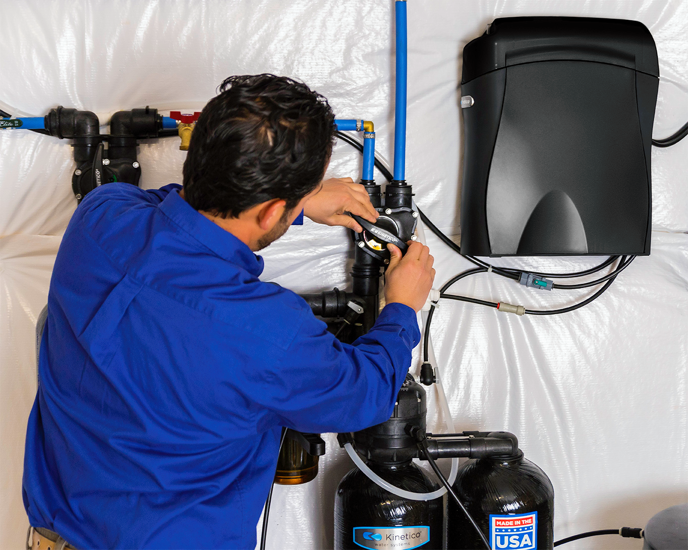 A person in a blue shirt installs or repairs a water softener system, connecting tubes and pipes against a white backdrop. Equipment and a control unit are mounted on the wall nearby.