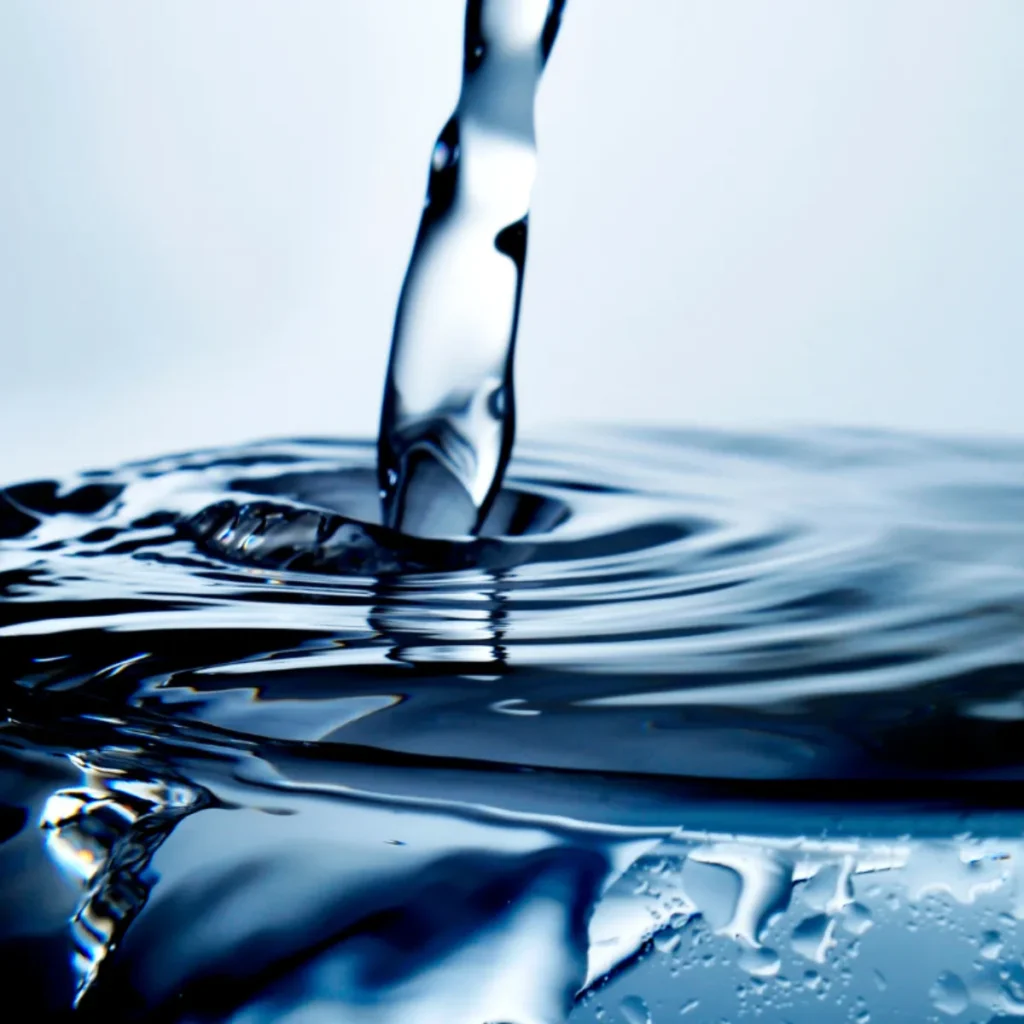 A close-up of a stream of water pouring into a calm, reflective pool, creating ripples and gentle waves on the surface. The image uses a blue tone, highlighting the clarity and movement of the water.