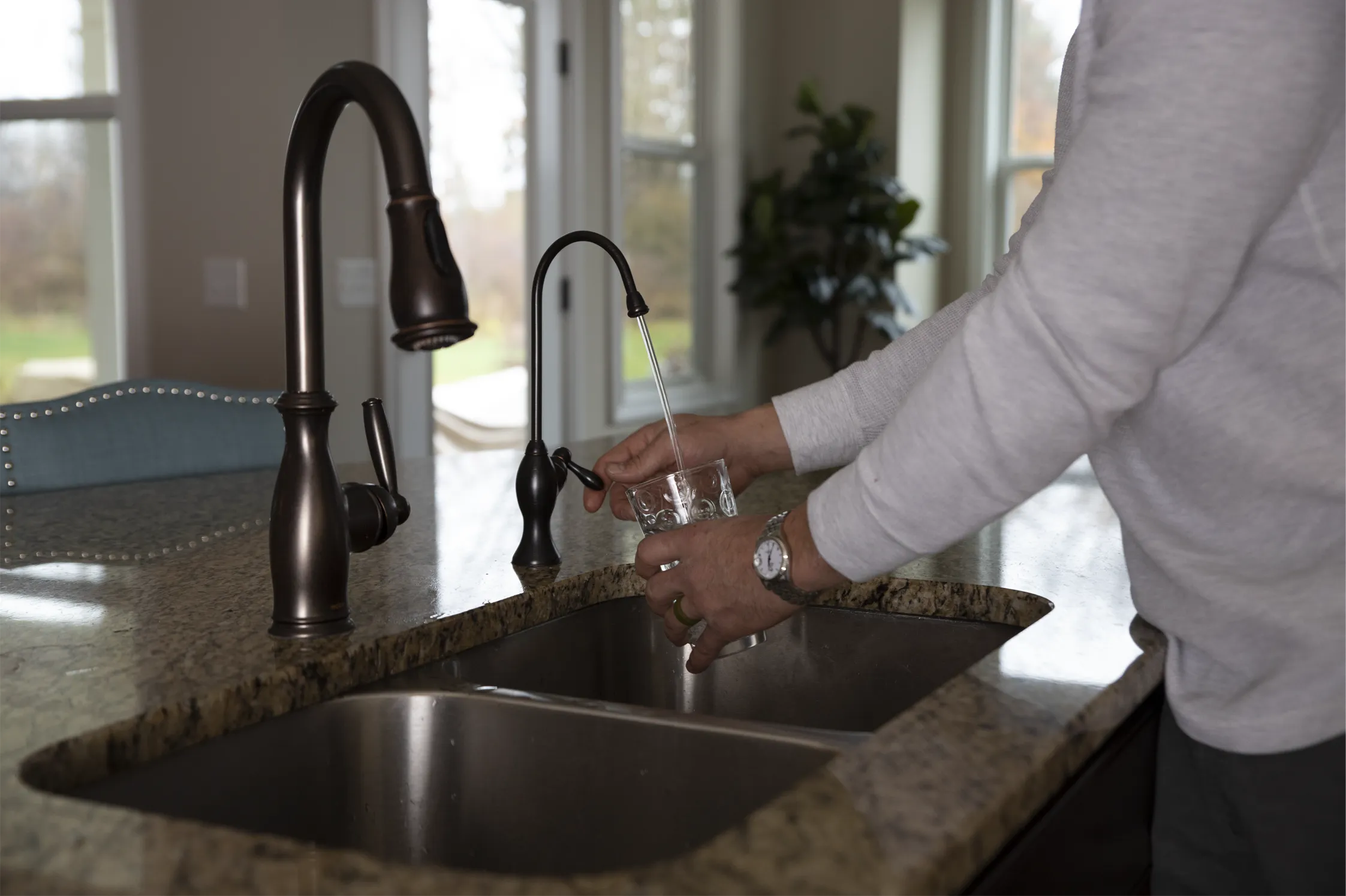 A person in a long-sleeve shirt fills a glass with water from a kitchen faucet, standing at a granite countertop and stainless steel sink in a well-lit room with large windows.