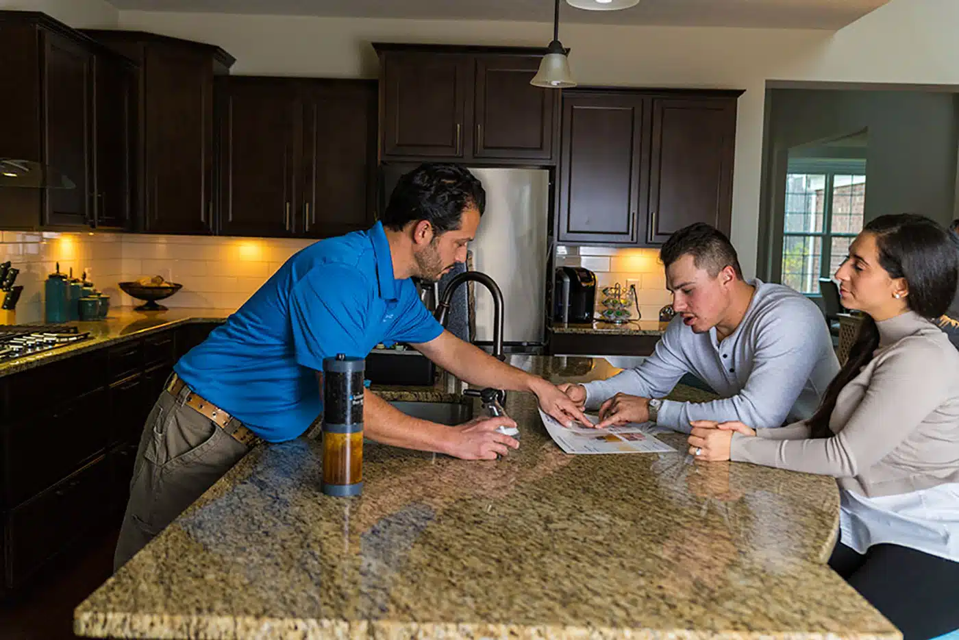 A man in a blue shirt explains documents to a seated couple at a kitchen counter. All three are focused on the paperwork, and the kitchen has dark cabinets and granite countertops.