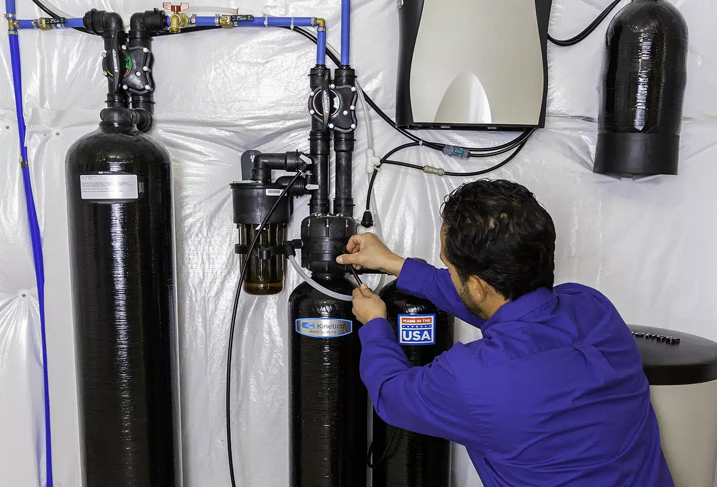 A man in a blue shirt installs or maintains a home water filtration system with large black tanks and pipes in a basement or utility room. The system has labels and an American flag sticker.