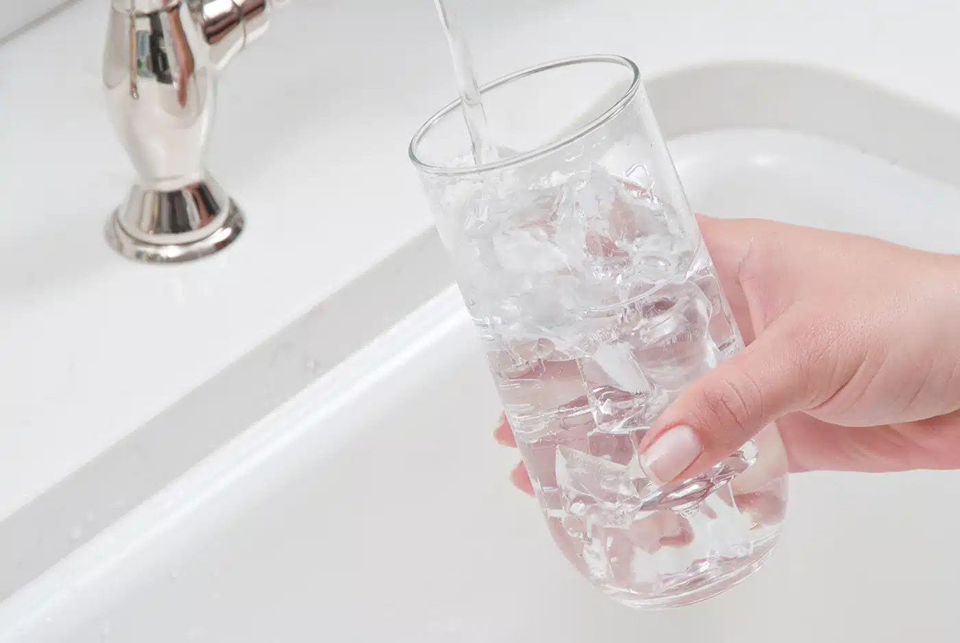 A hand holds a glass filled with ice under a faucet, as water is being poured into the glass over a white sink.