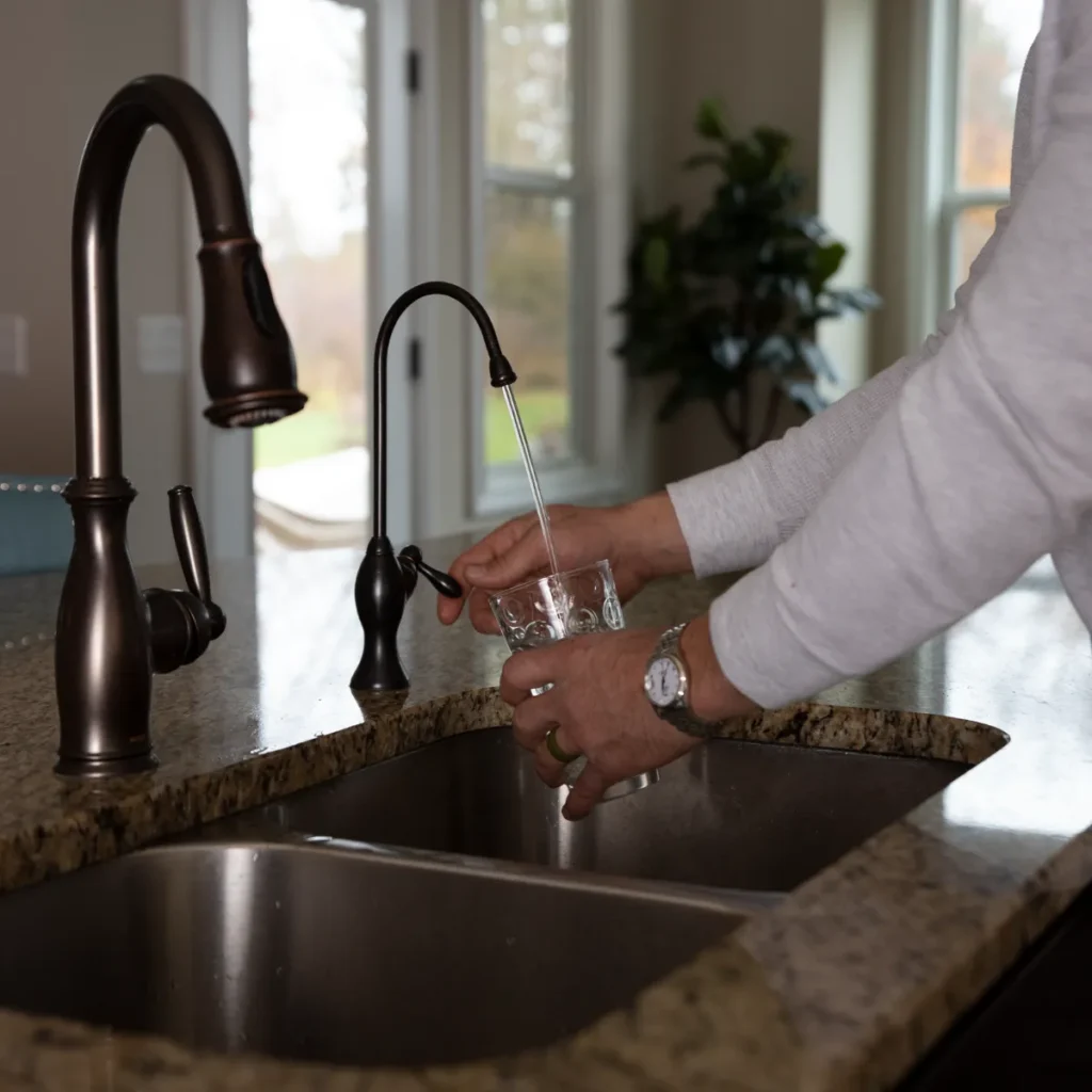 A person fills a glass with water from a small faucet on a granite kitchen countertop, next to a larger sink faucet, with a plant and windows in the background.