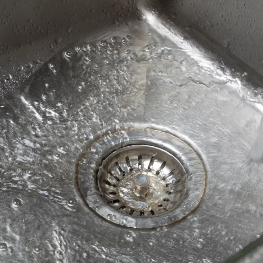 Close-up of water spinning and draining in a stainless steel kitchen sink, with droplets and reflections visible on the metal surface.