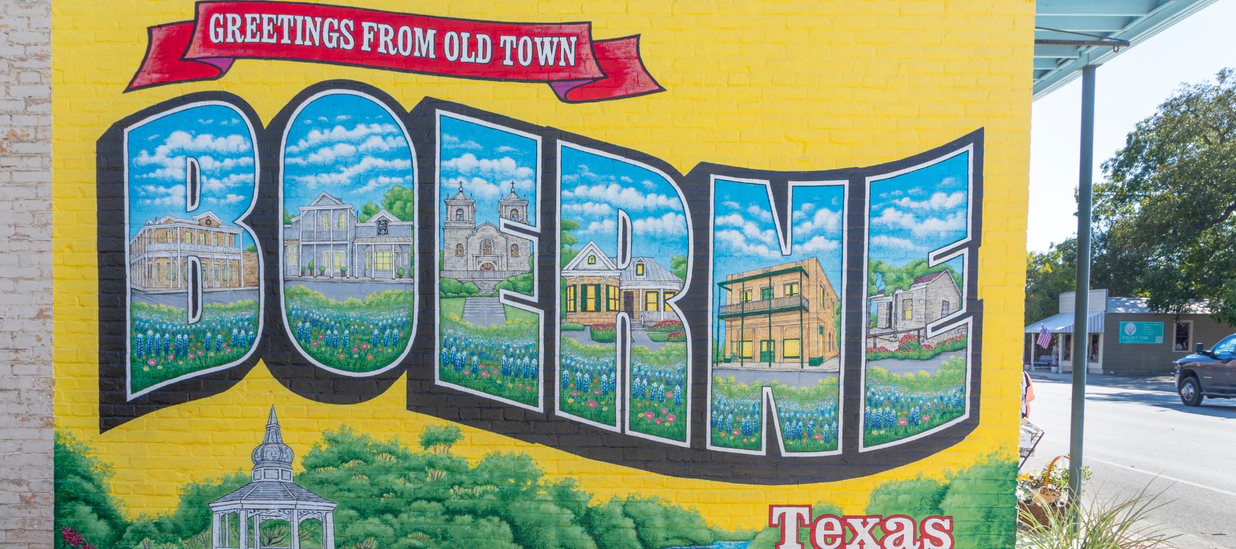 A mural on a yellow brick wall reads “Greetings from Old Town Boerne, Texas,” with large letters filled with images of local buildings, wildflowers, and a white gazebo at the bottom.