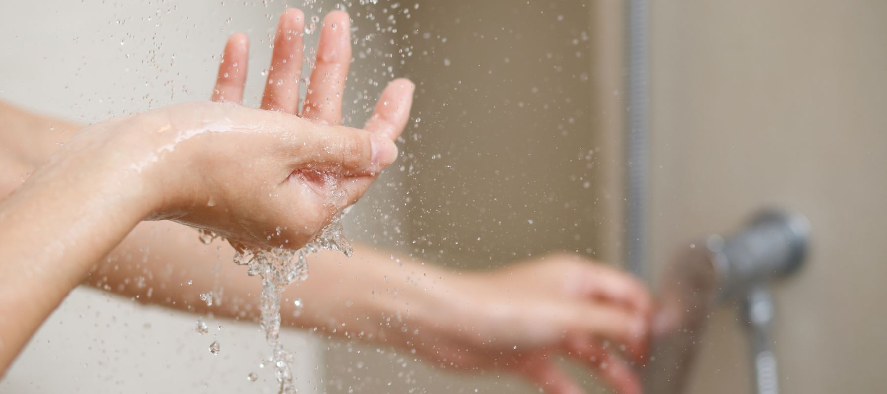 A close-up of two hands under running water, with droplets splashing, possibly in a shower or near a sink. The background is blurred, focusing attention on the hands and the flowing water.