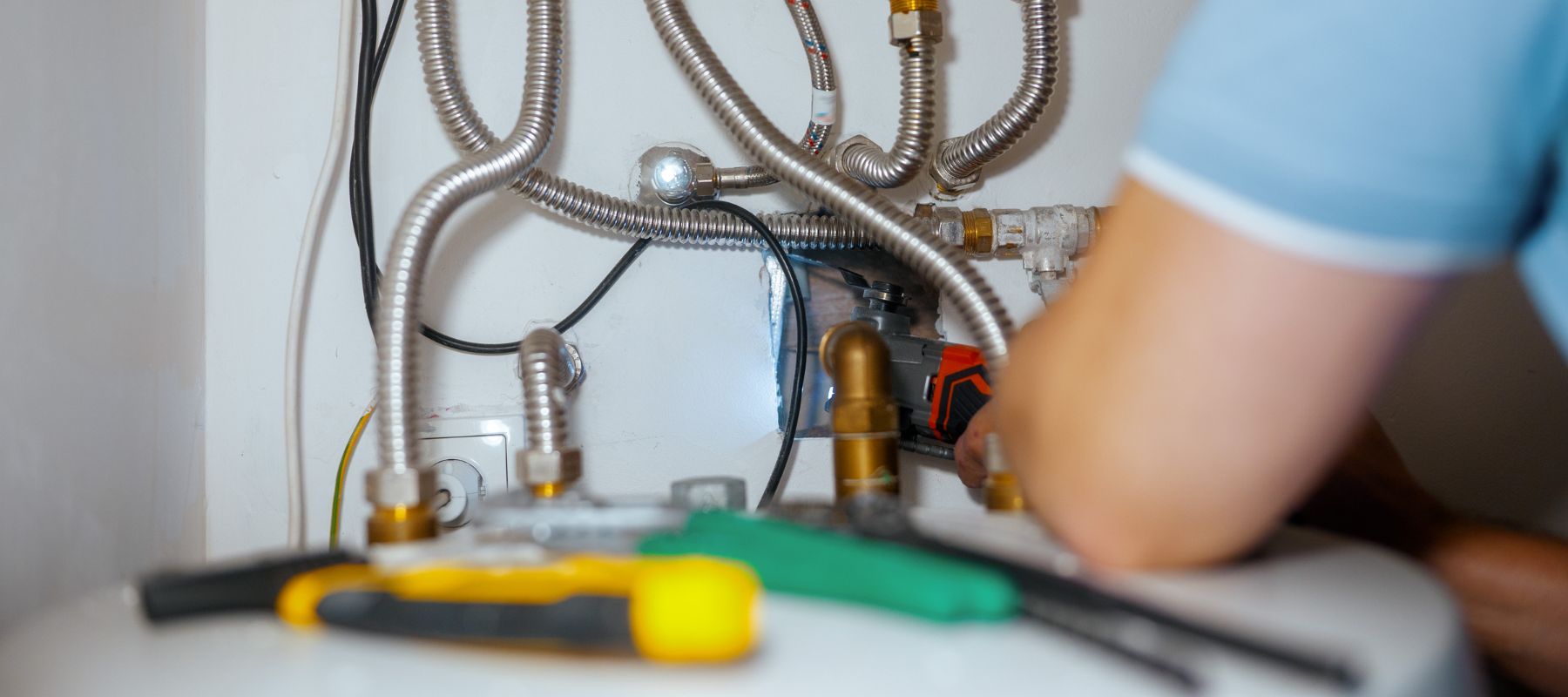 A person is working on plumbing connections, surrounded by metal flexible pipes and valves. Tools including pliers and a screwdriver are placed in the foreground. Only the worker’s arm and shoulder are visible.