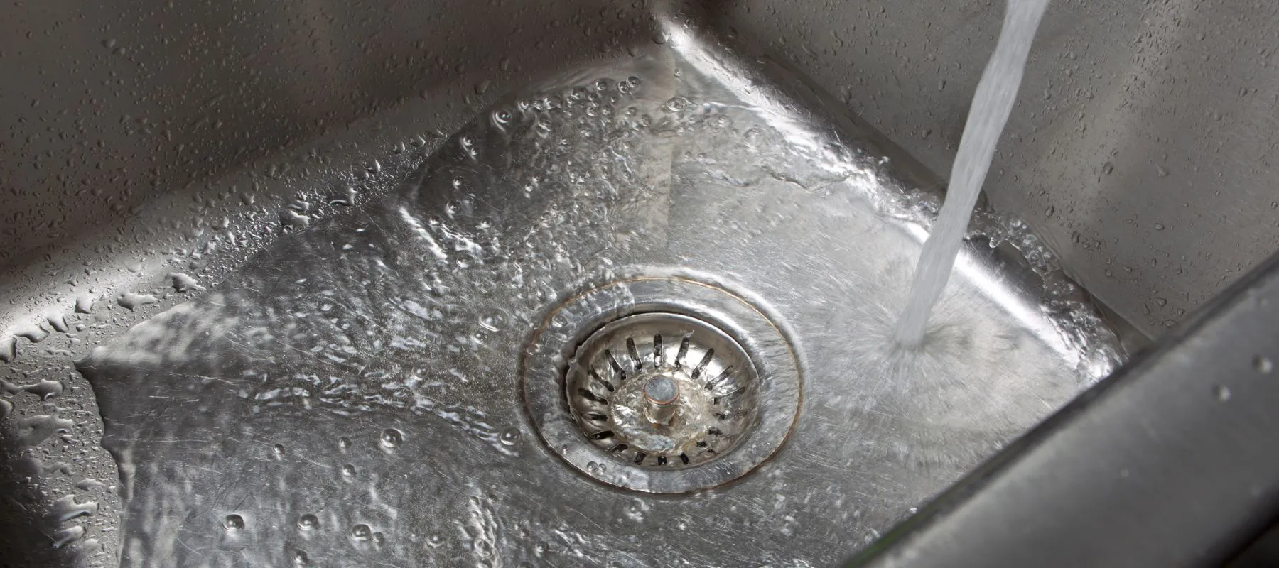 A stream of water flows from a faucet into a stainless steel sink, surrounding the drain and creating ripples and reflections on the metal surface.