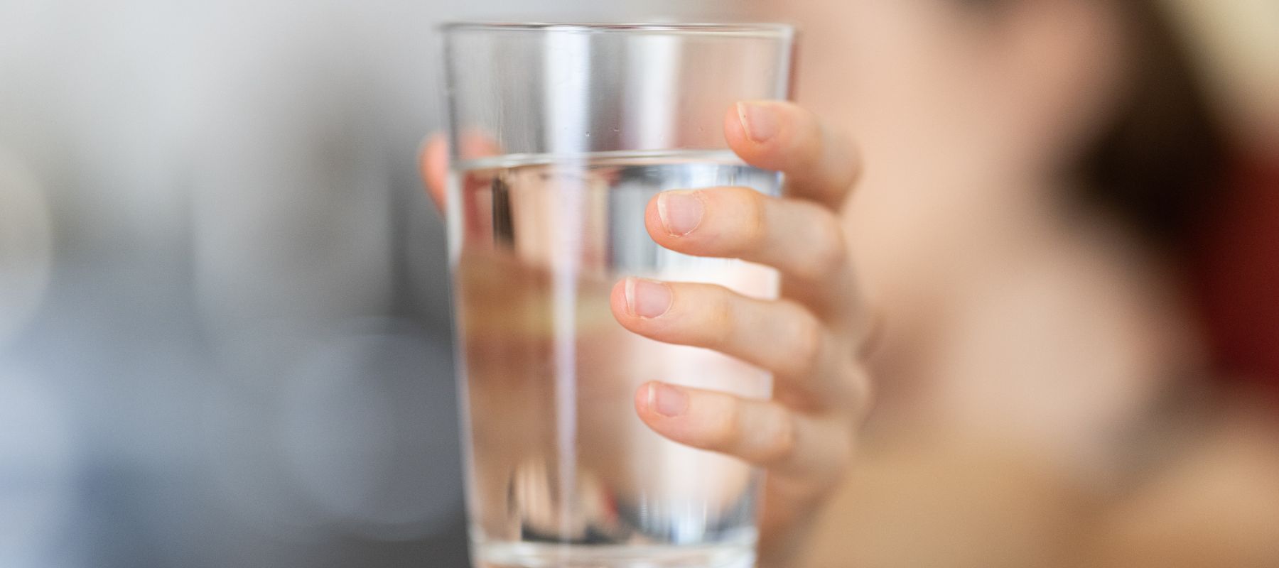 A person holding a clear glass of water, with their hand and fingers visible in focus and the background blurred.