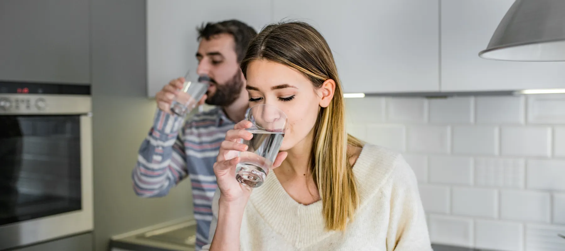 A woman and a man stand in a modern kitchen, both drinking water from clear glasses. The woman is in the foreground, wearing a light sweater, while the man is in the background in a striped shirt.