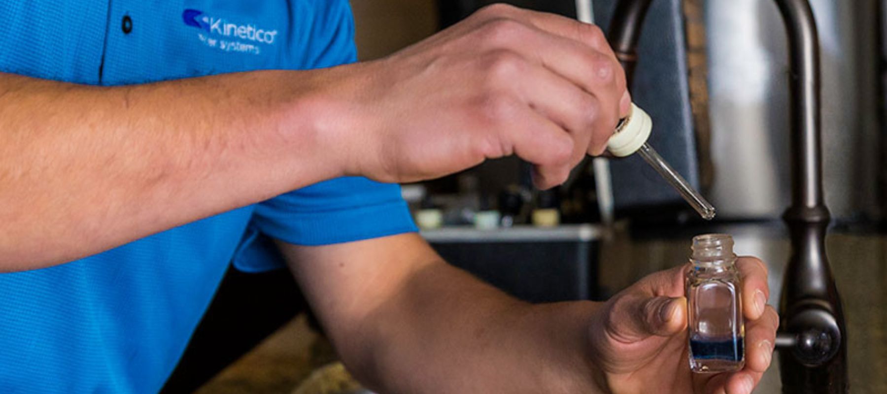A person in a blue shirt uses a dropper to add liquid to a small glass vial, possibly conducting a water test near a faucet.
