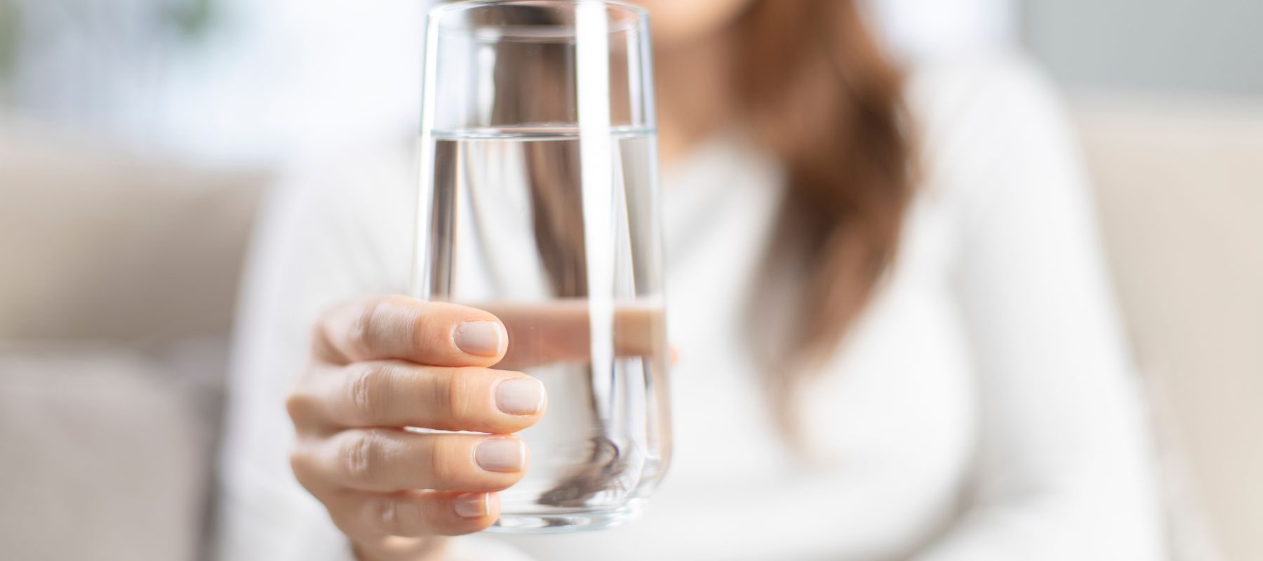 A person with long brown hair, wearing a white top, holds a clear glass of water toward the camera. The background is softly blurred, focusing on the hand and the glass.