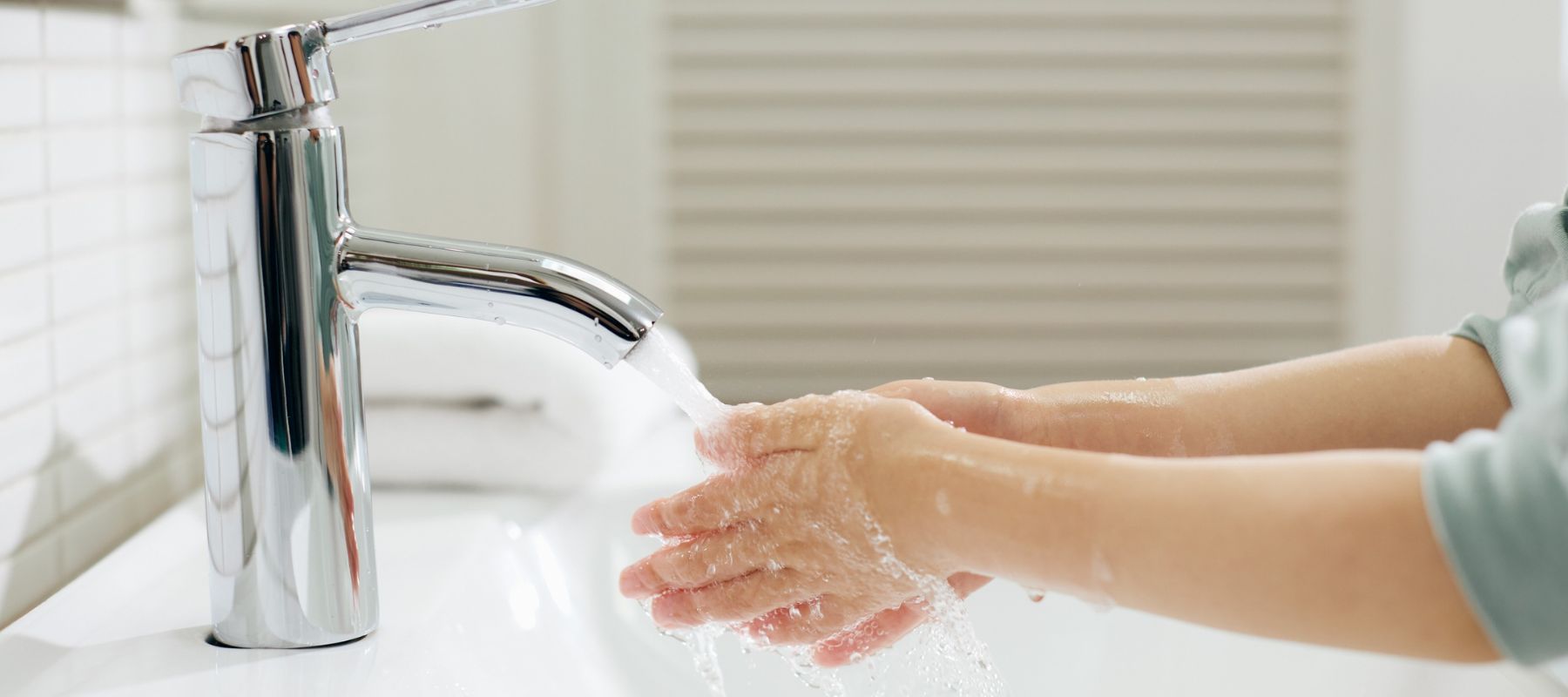 A person washing their hands under running water from a modern, silver faucet at a white sink, with soap and bubbles visible on their hands.