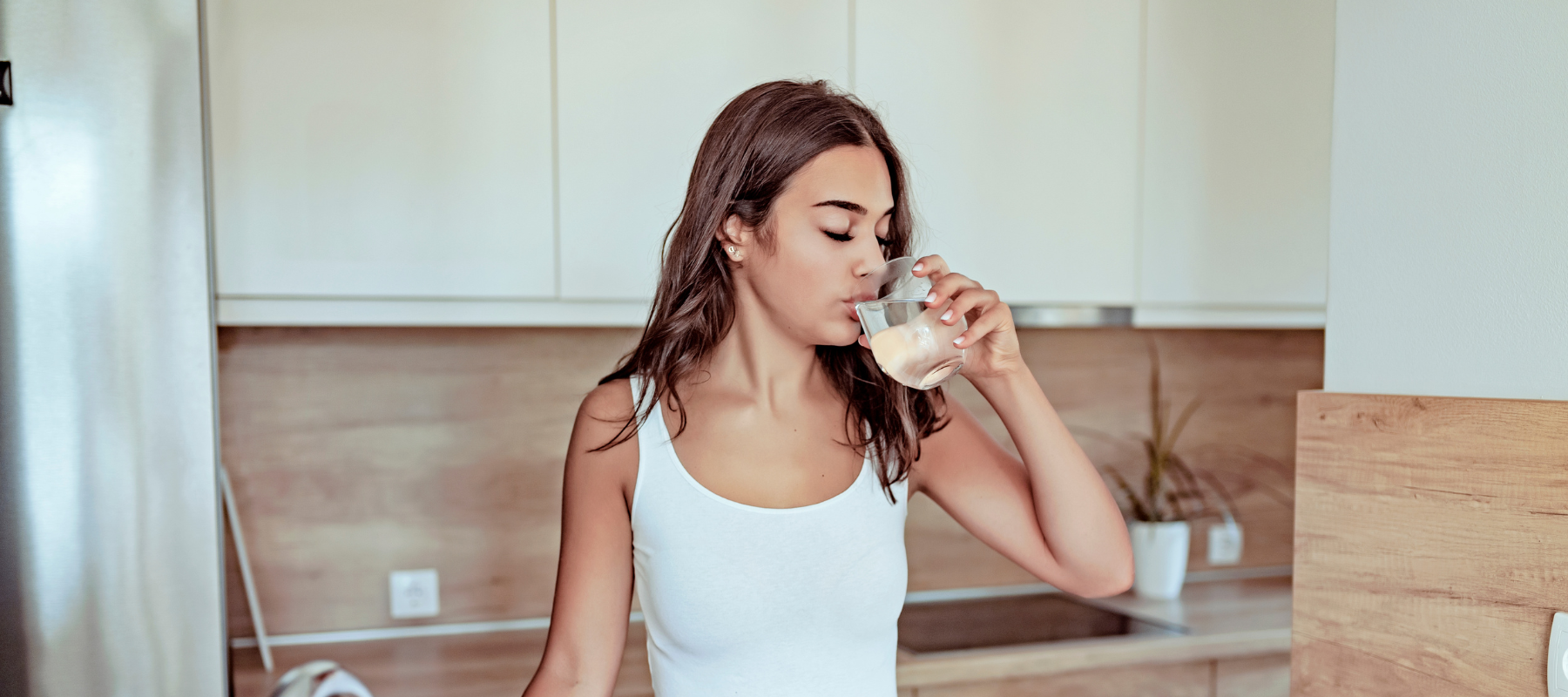 A young woman with long brown hair, wearing a white tank top, stands in a modern kitchen and drinks from a glass.