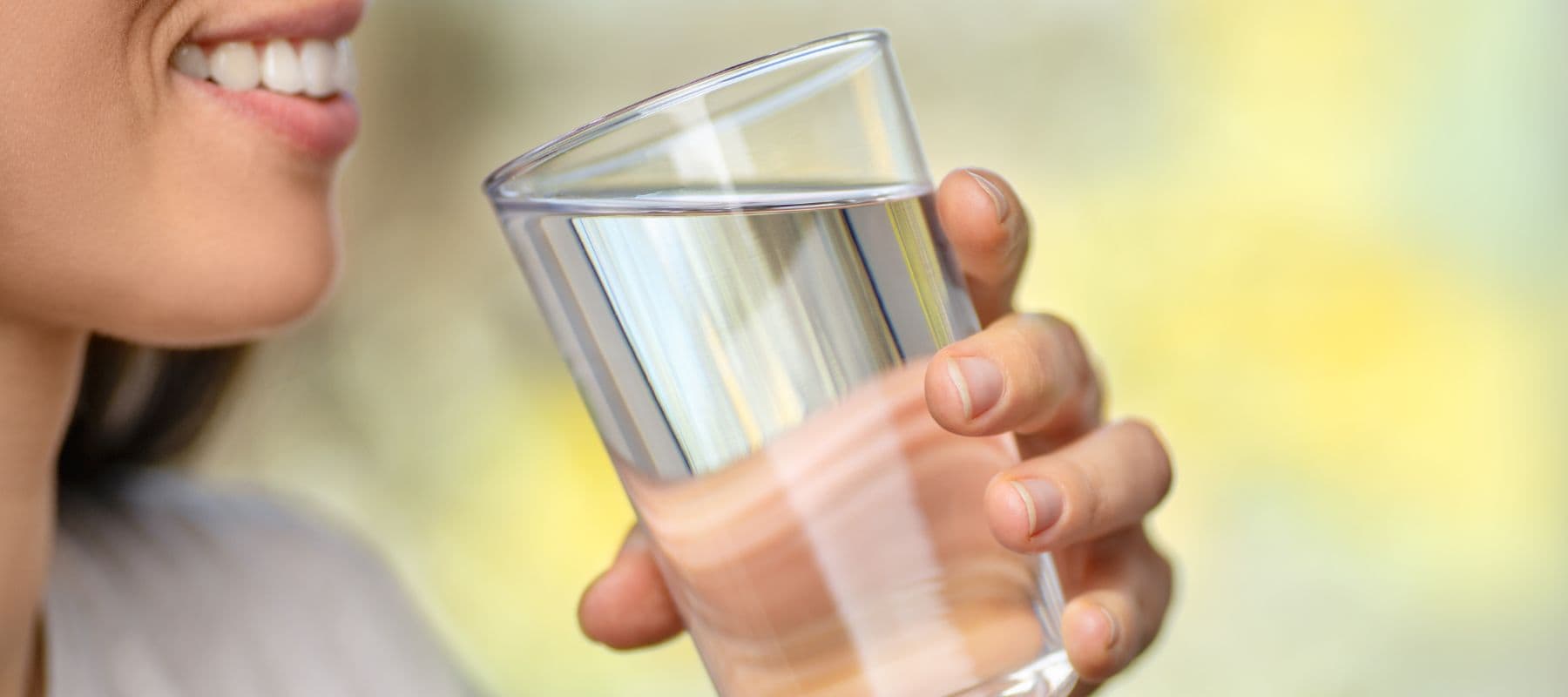 A person smiling and holding a clear glass of water close to their mouth, ready to take a sip. The background is blurred with light green and yellow tones.