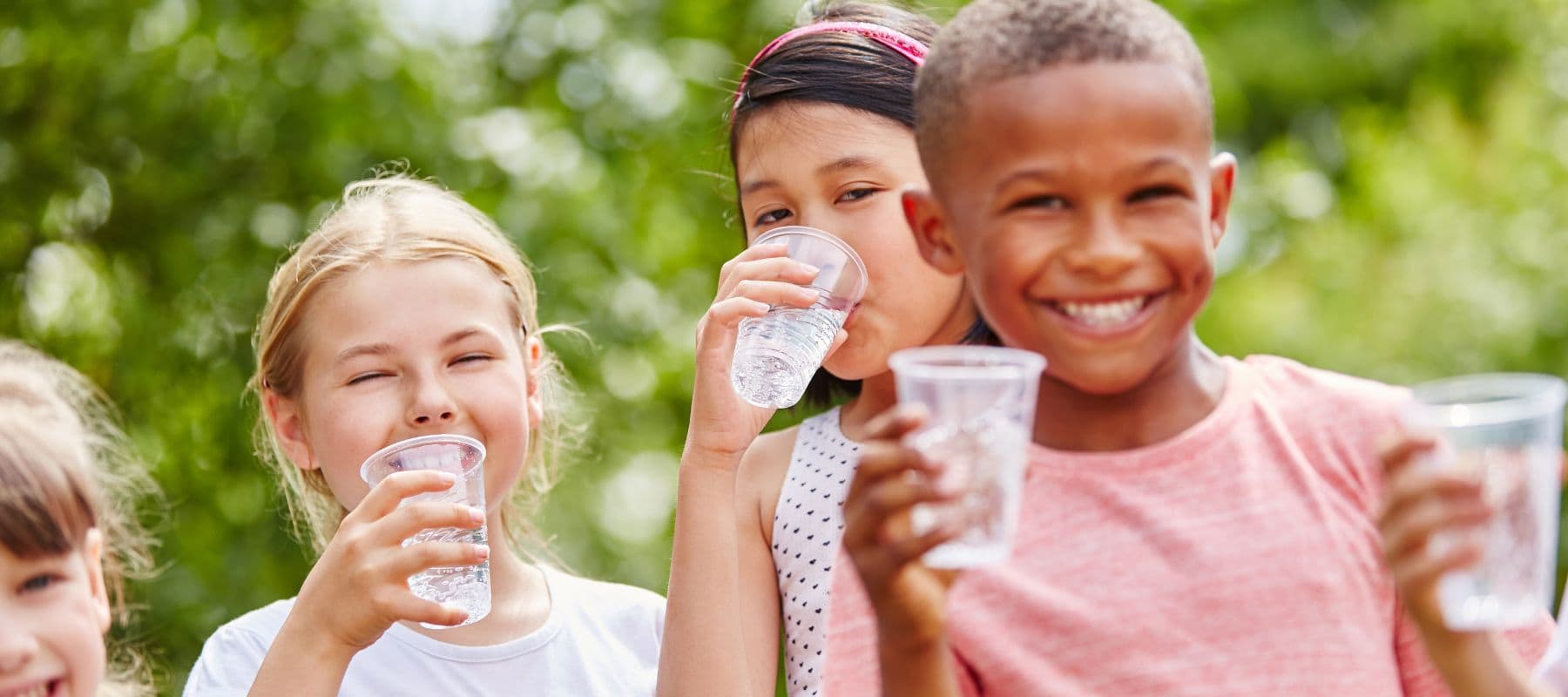 Four children stand outdoors, smiling and holding clear plastic cups of water. Two girls are drinking, while a boy in front smiles brightly at the camera. Green trees are blurred in the background.