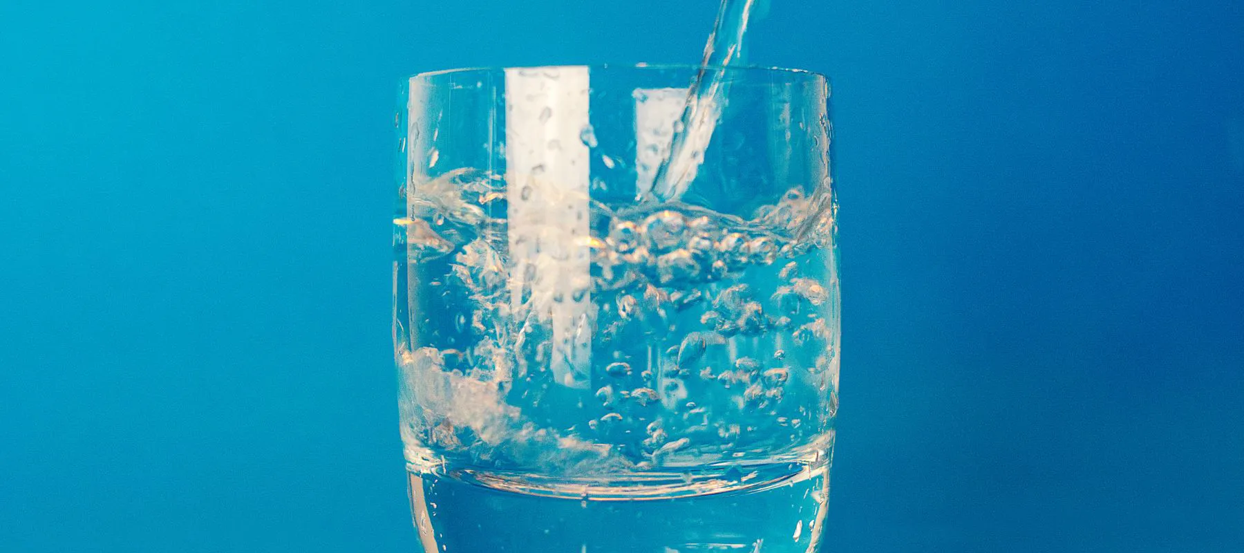 A close-up of water being poured into a clear glass, creating bubbles, with a bright blue background.