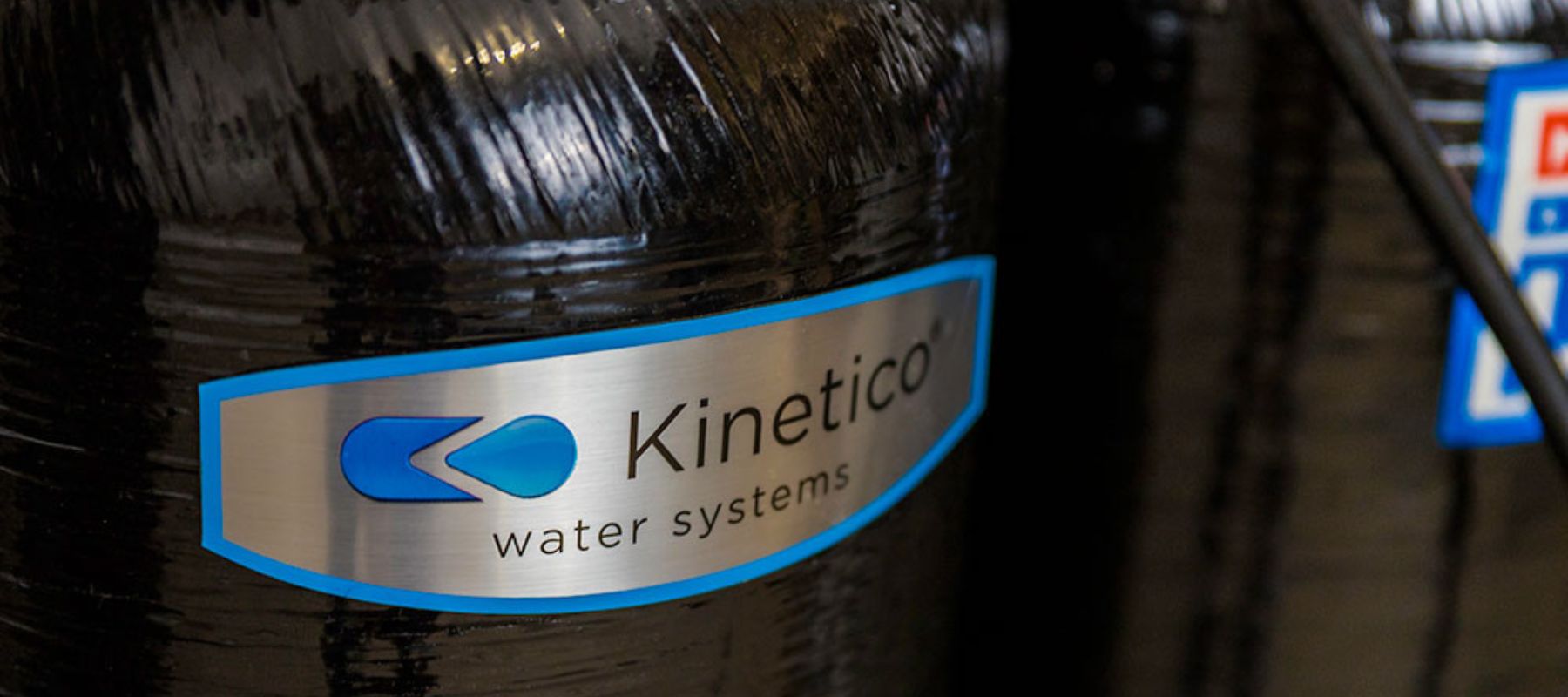 Close-up of a black water filtration tank with a metallic label that reads Kinetico water systems in black letters and a blue logo on the left side.