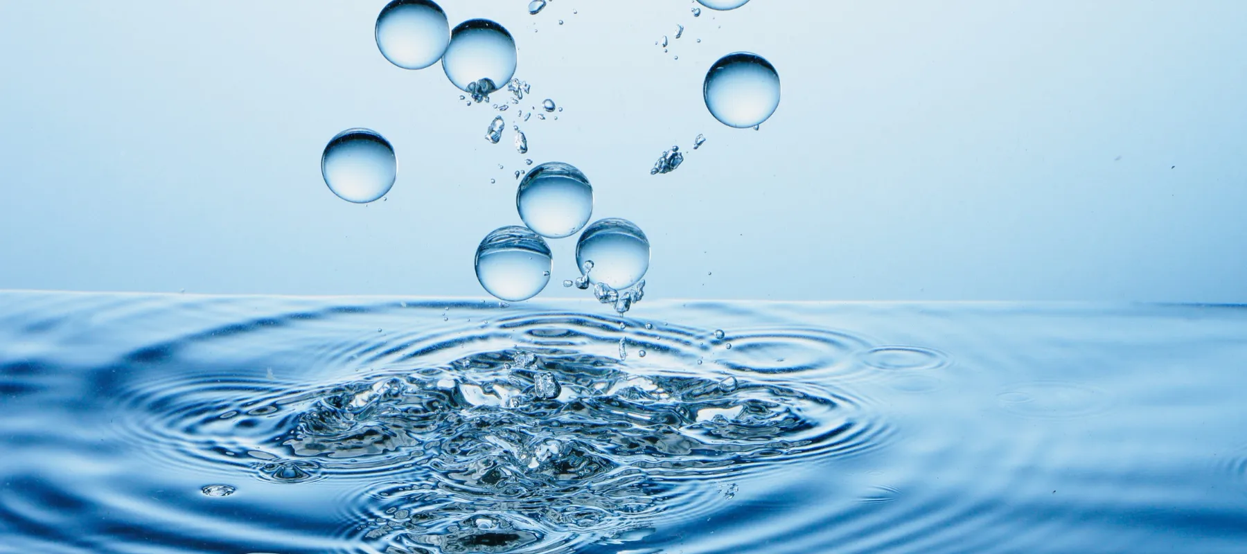Clear water droplets falling and splashing into a blue water surface, creating ripples and bubbles with a smooth, gradient blue background.
