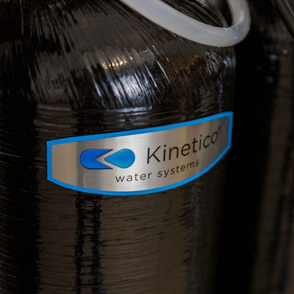 Close-up of a black tank labeled Kinetico water systems with a blue logo, part of a water filtration or softening system. A white tube is attached at the top.