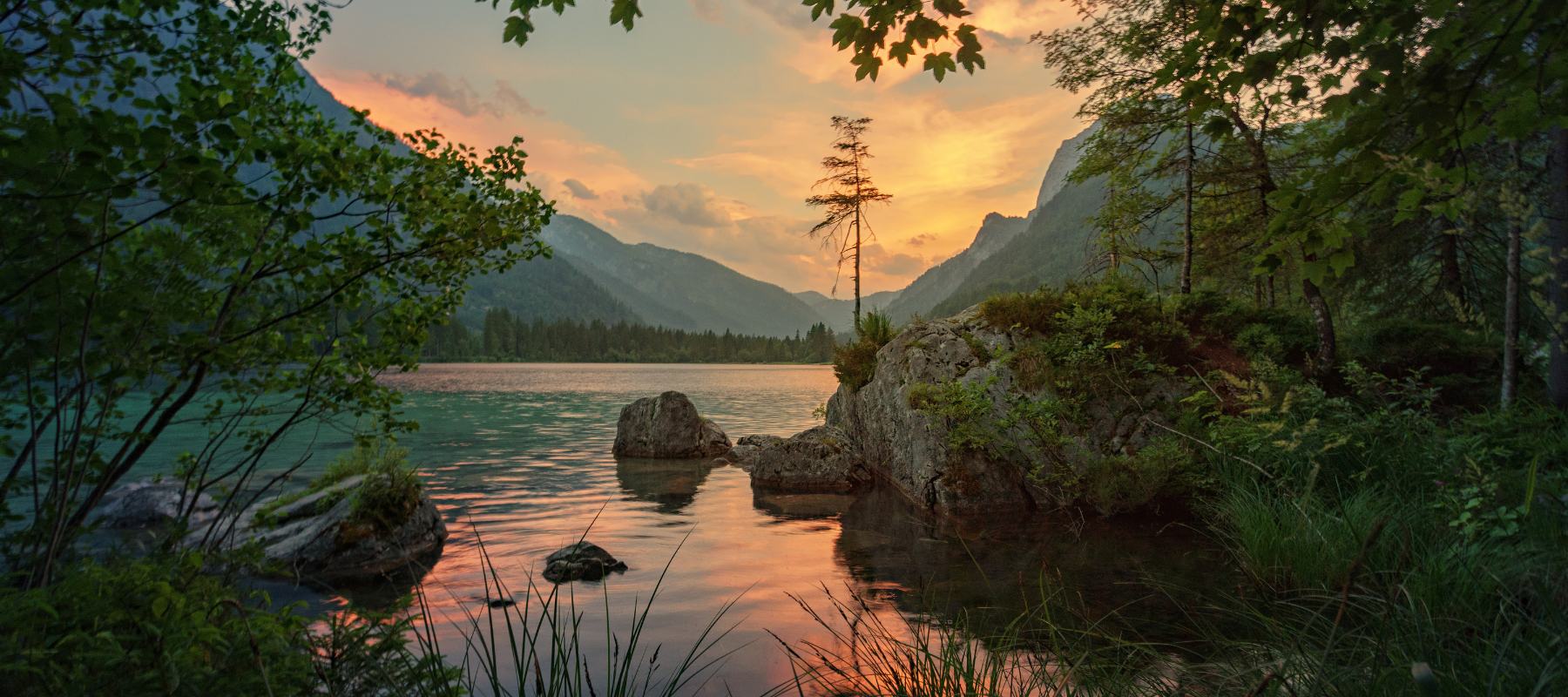 A serene lake surrounded by lush greenery and mountains at sunset, with trees and rocks in the foreground and colorful clouds reflecting on the calm water.