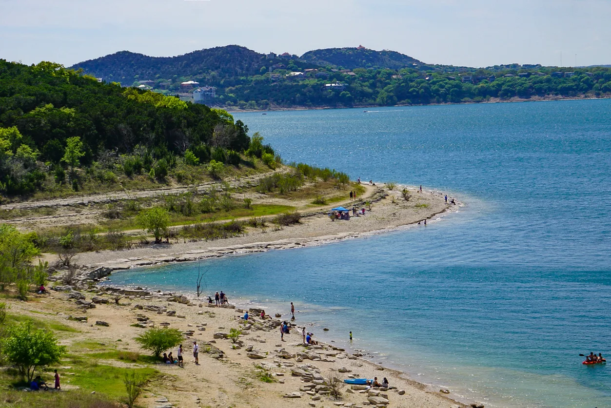 A curved shoreline along a lake with people scattered on the rocky beach, some near the water and others under shade. Green hills and houses are visible in the background across the lake.
