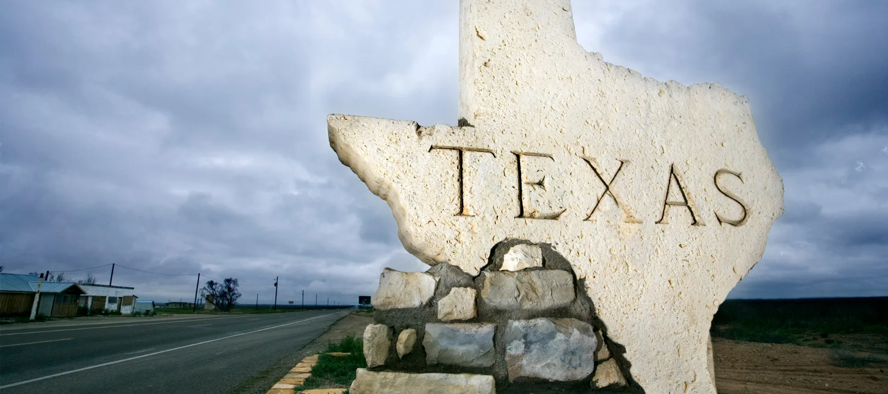 A large stone monument shaped like the state of Texas, engraved with the word TEXAS, stands beside a rural road under a cloudy sky.