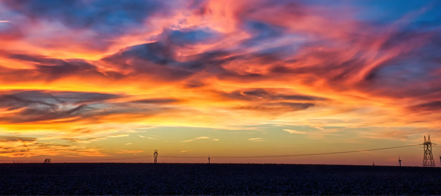 Vivid sunset with dramatic clouds in shades of orange, pink, and purple above a dark, flat landscape. Silhouettes of utility poles and wires are visible against the colorful sky.